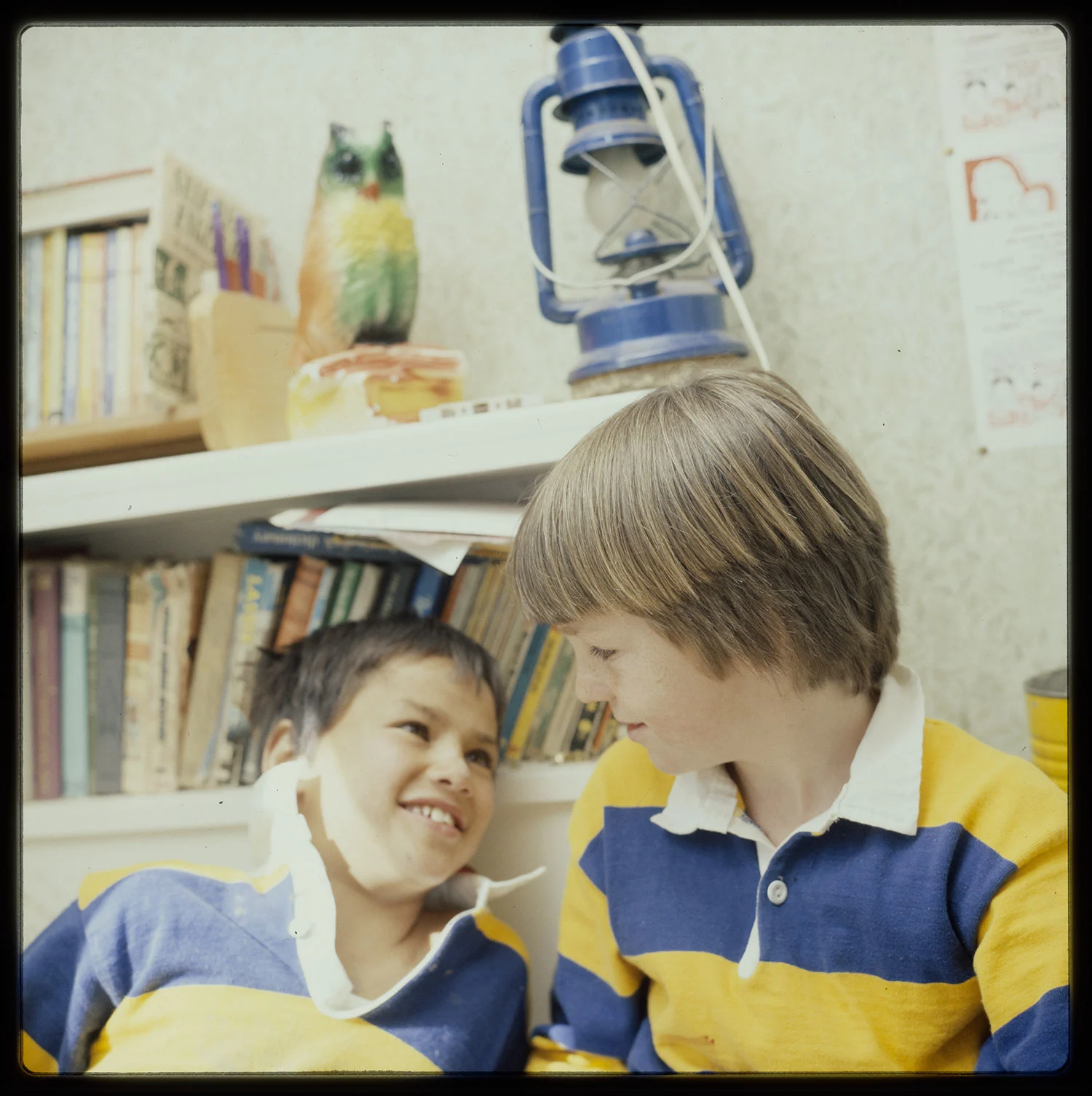 Two boys wearing a yellow and blue striped t shirt and shorts sitting on a bed, a book case with books at the back