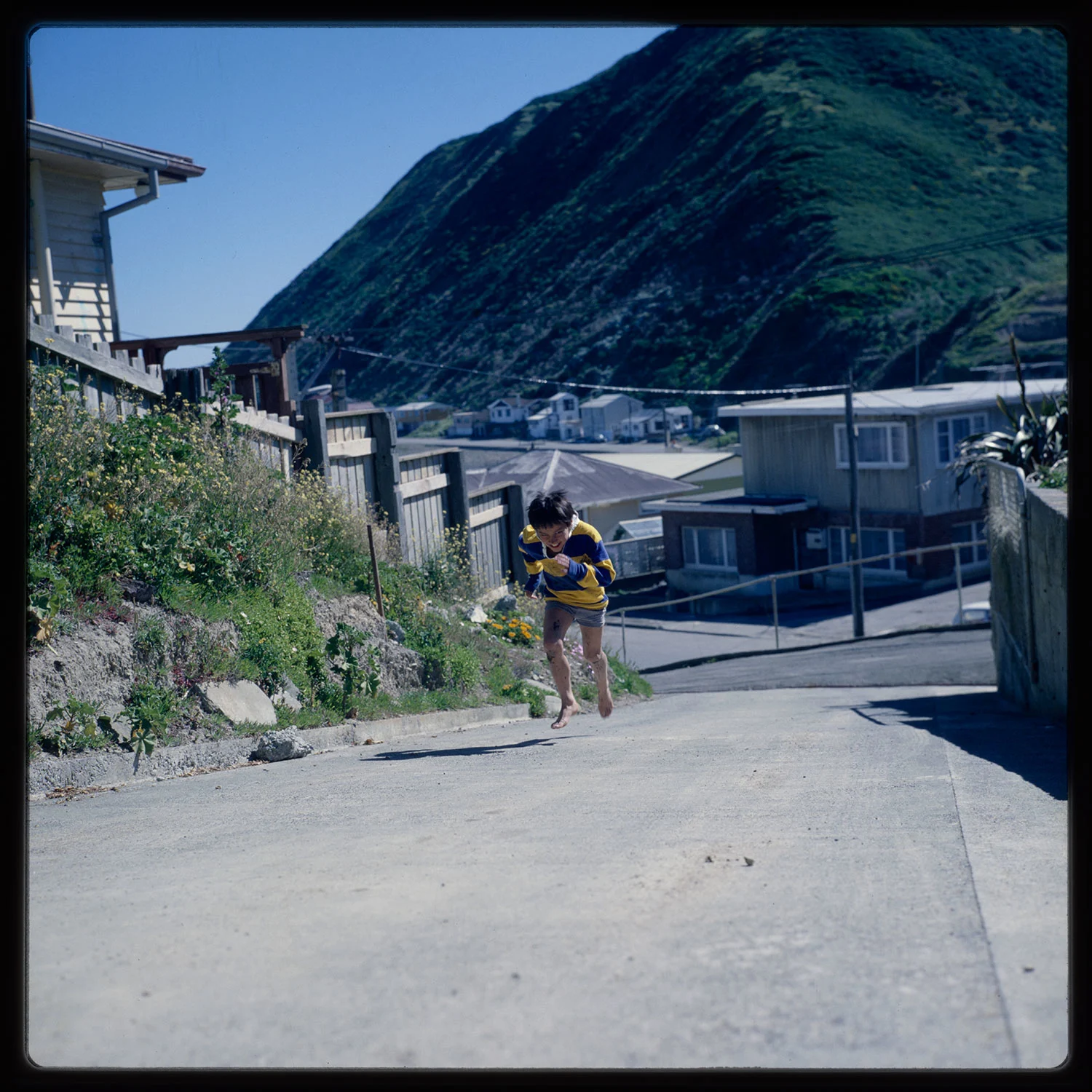 A boy wearing a yellow and blue striped shirt running on an uphill road with houses in the background