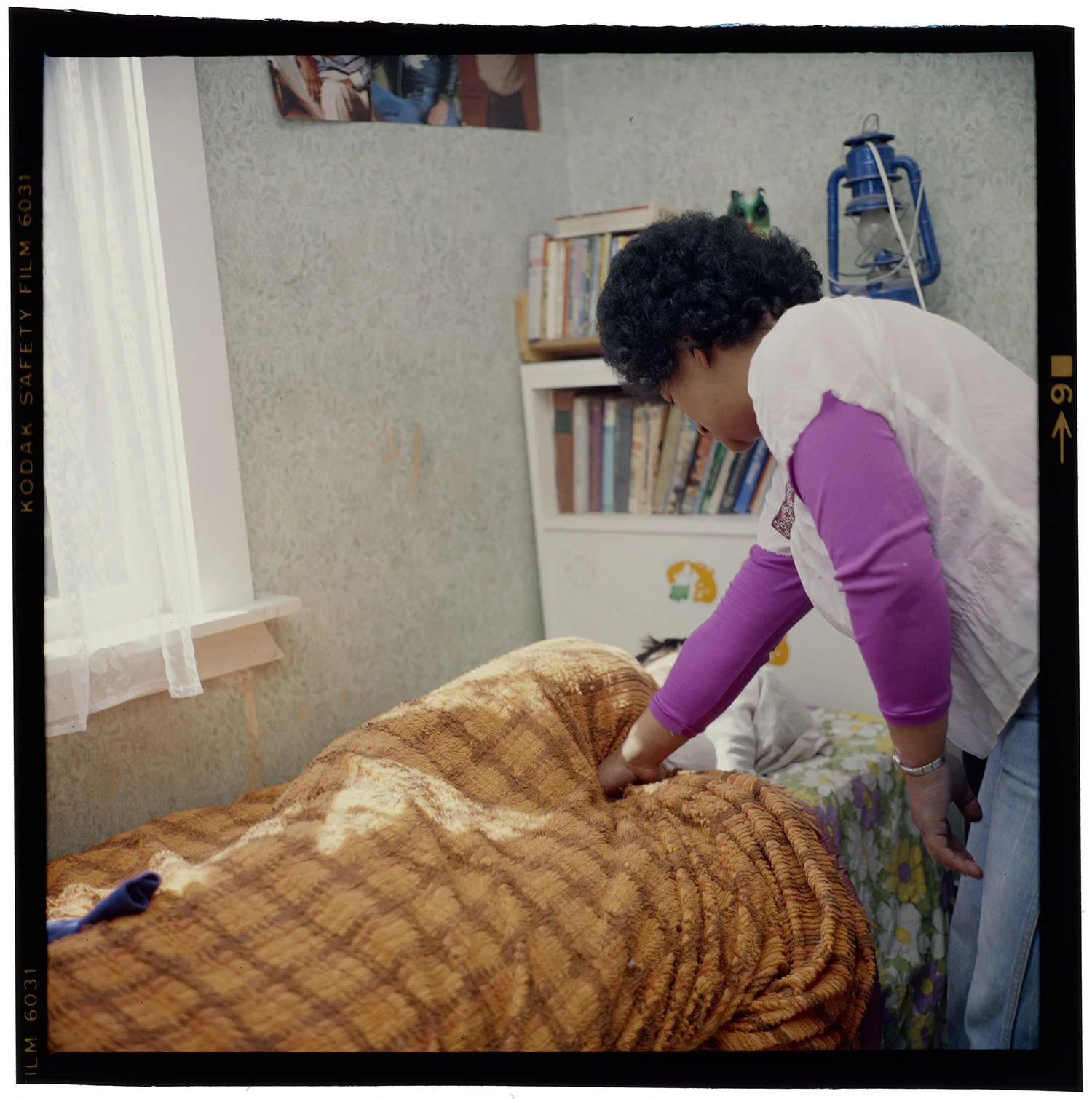 A woman with short hair wearing a purple long sleeves shirt and a white top trying to wake up a boy sleeping on a bed