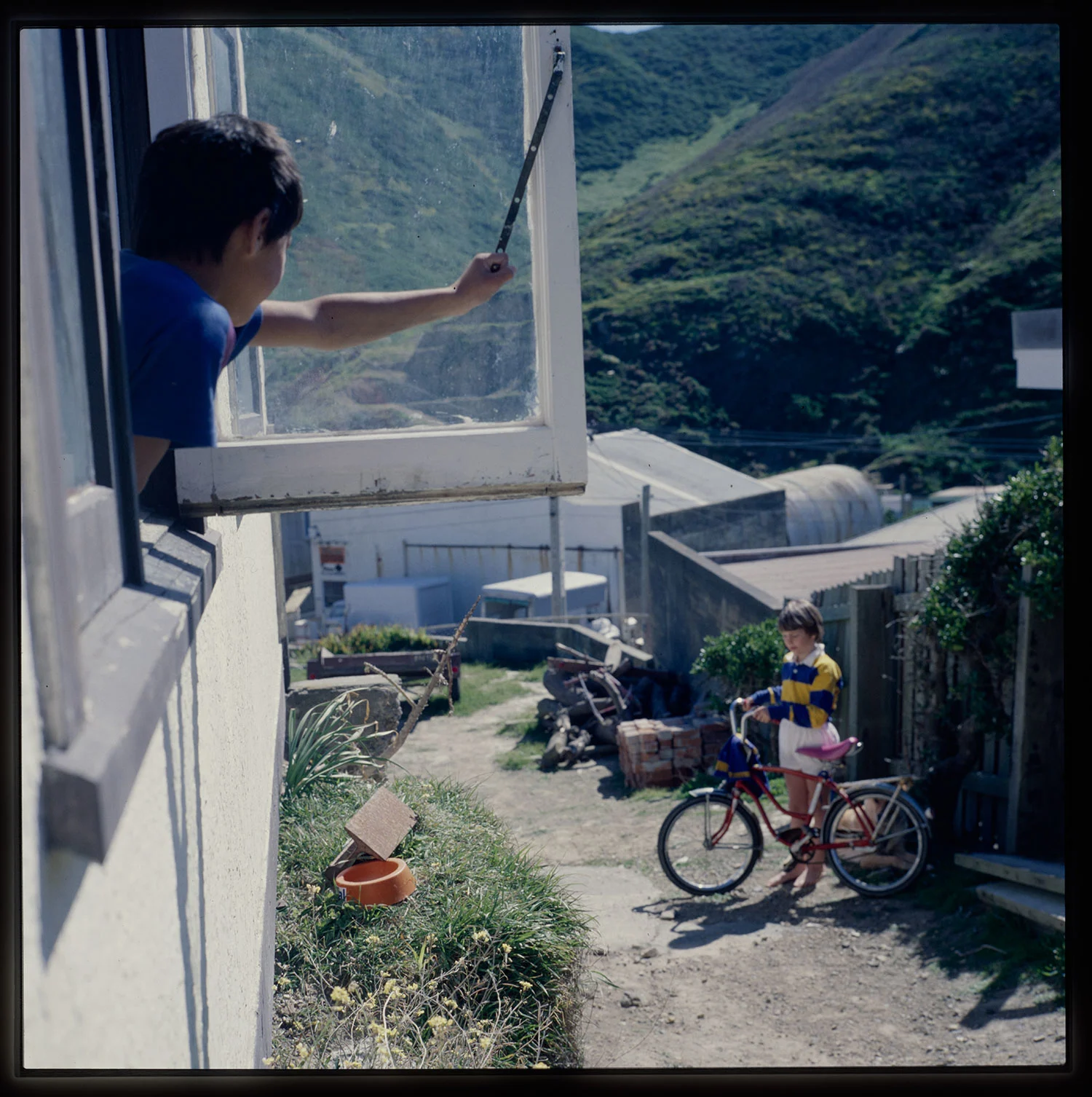 A boy peeping out of an open glass window, another boy standing outside holding a bike in his hands