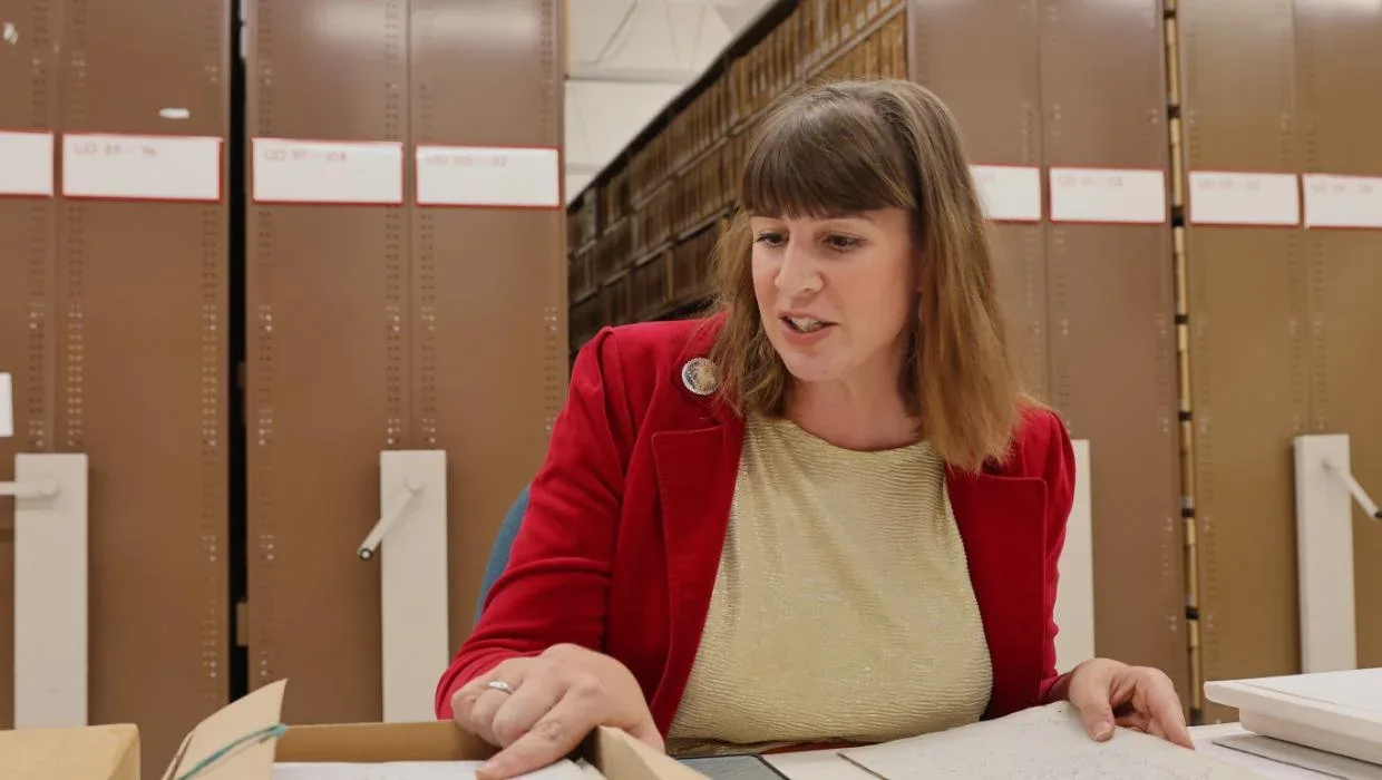 Woman with mustard t-shirt and red jacket sitting with a box and papers in front of her.