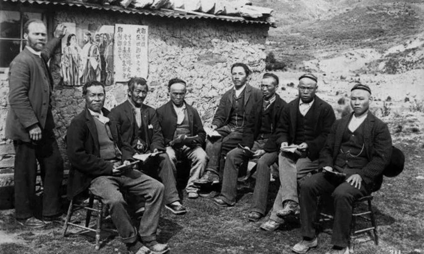 Black and white photo of seven men sitting on chairs and one man on the left standing, an old hut in the background 