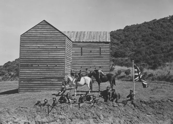 Soldiers prepare to fire rifles alongside flag. Wooden fortress behind them and two horses with riders.