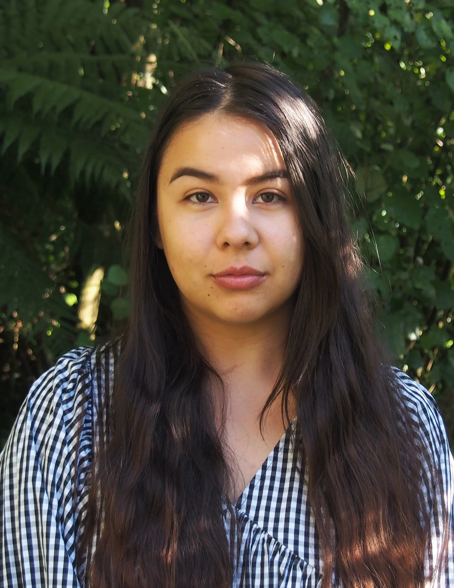 A woman with long hair and wearing a frock, standing in front of green trees with sunlight on her face