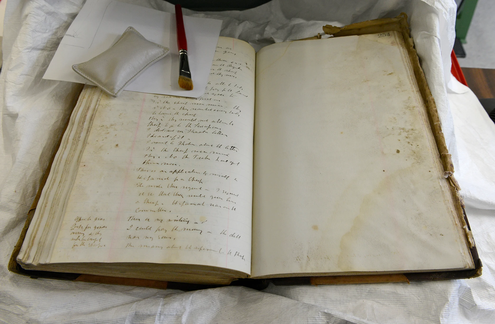 An open aged book with handwritten text on its pages is laid on a table. The book's pages show visible signs of age and wear, and there is another piece of paper resting on the book with a brush and small cushion.