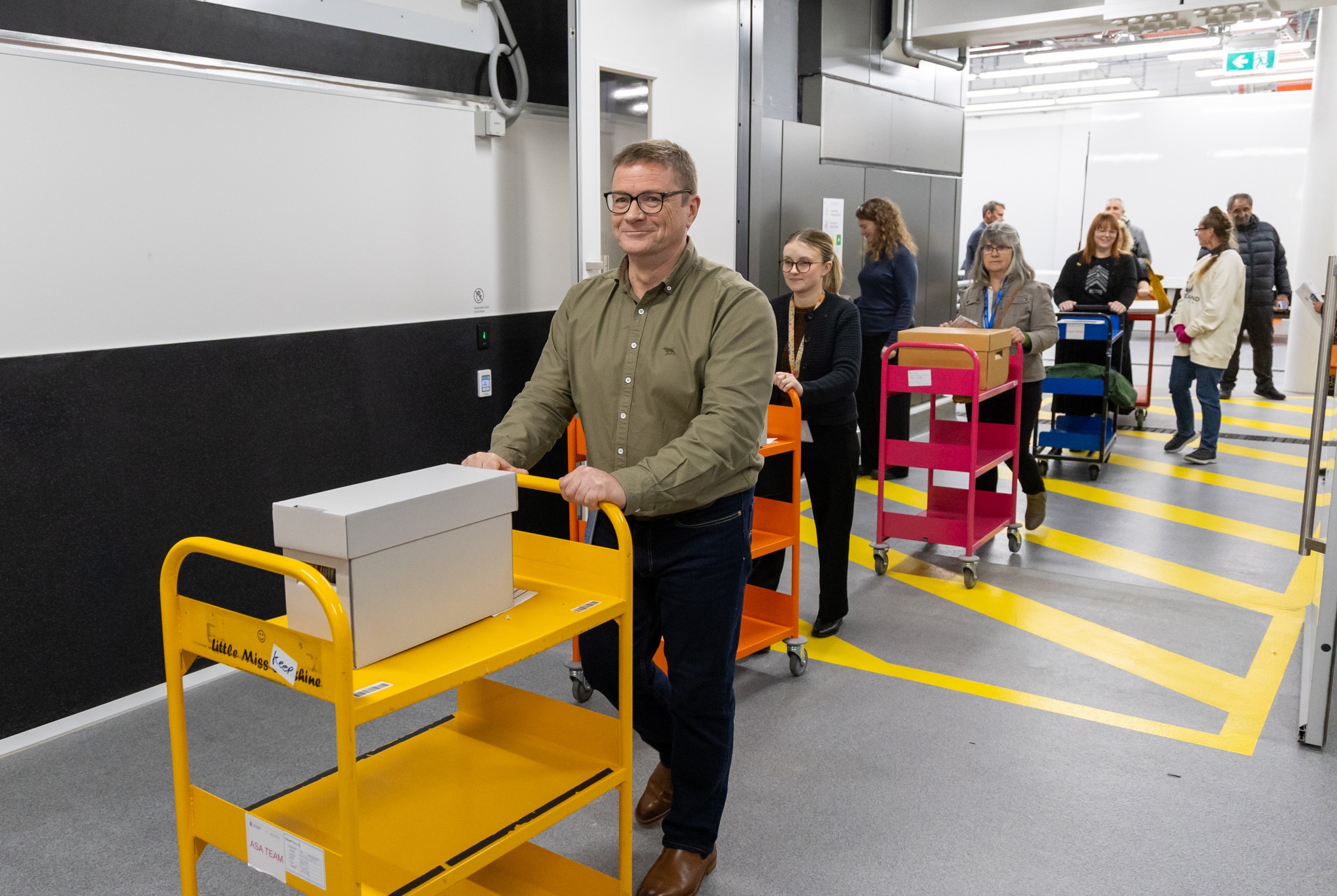 A man wearing glasses, green shirt and black pant is pushing a yellow trolley. A box is kept on the trolley.