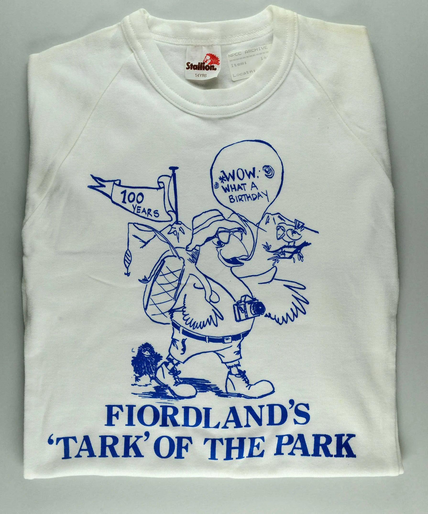 A folded beige coloured shirt with a blue cartoon and words written 'Fiordland's 'Tark' of the park.  