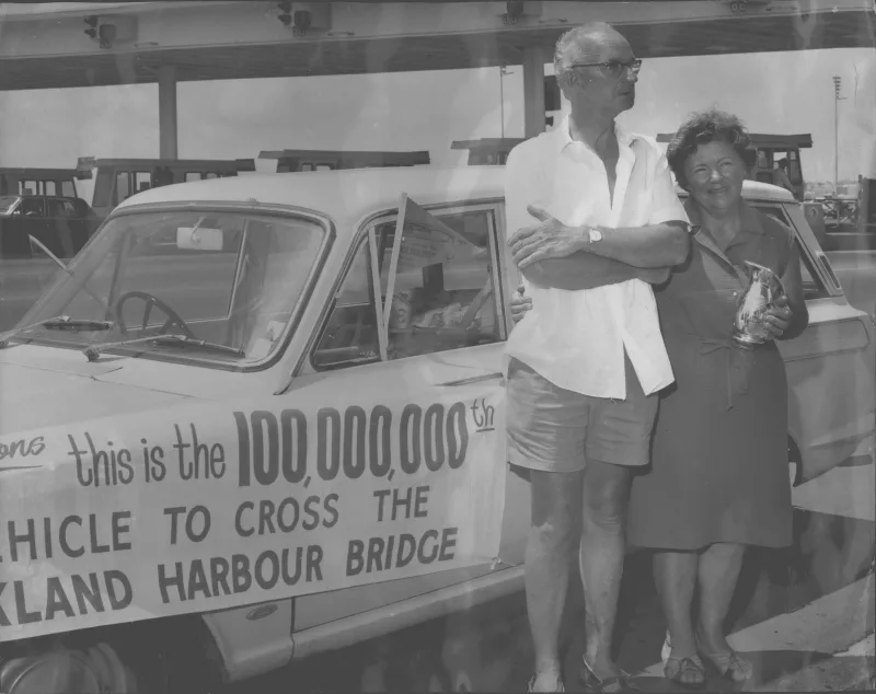 Black and white photo showing a man and a women standing in front of a car with a banner announcing "This is the 100,00,000 vehicle to cross the Auckland Barbour Bridge"