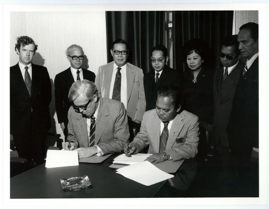 2 men sit at a table signing documents while 7 others stand around them.