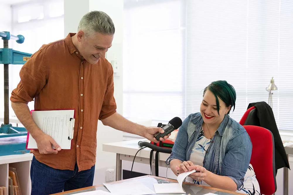 Photograph of a Gareth Watkins holding a microphone up to Lenette Breytenbach,  Royal Commission Digitisation Assistant