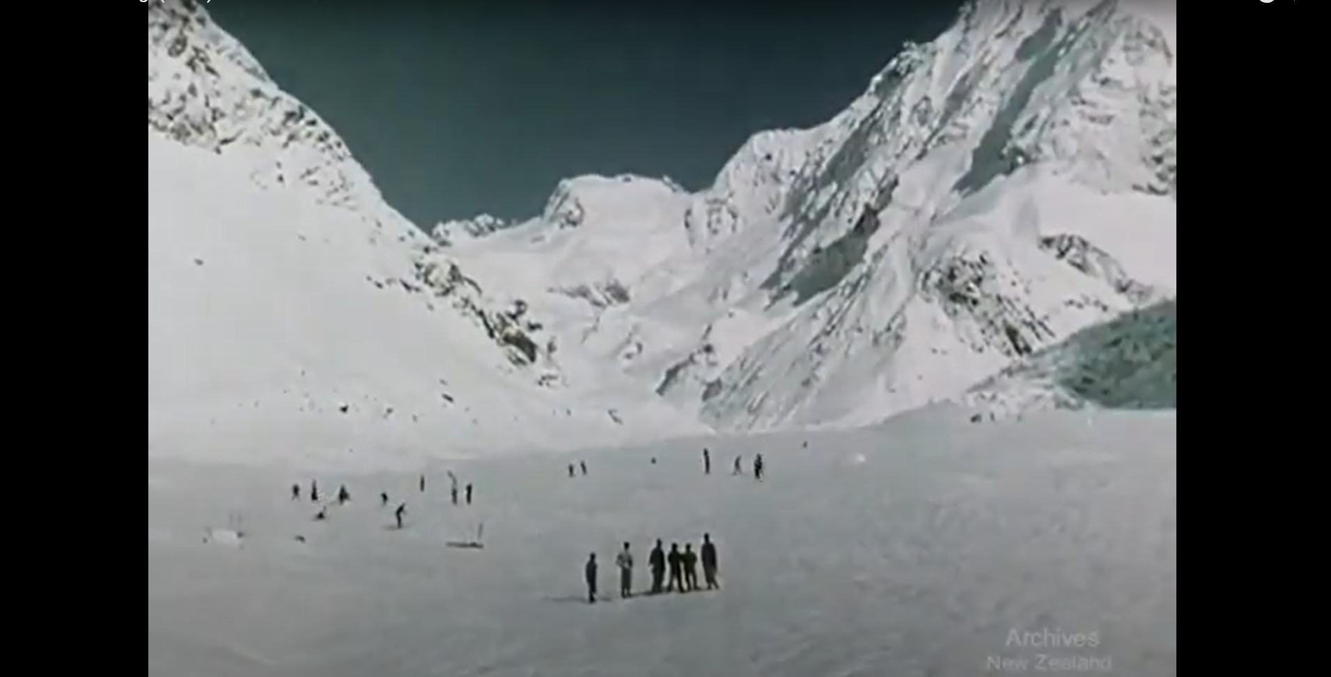 An old photo of white mountains on a background of dark green sky, with few people standing in the foreground and some people skiing.  