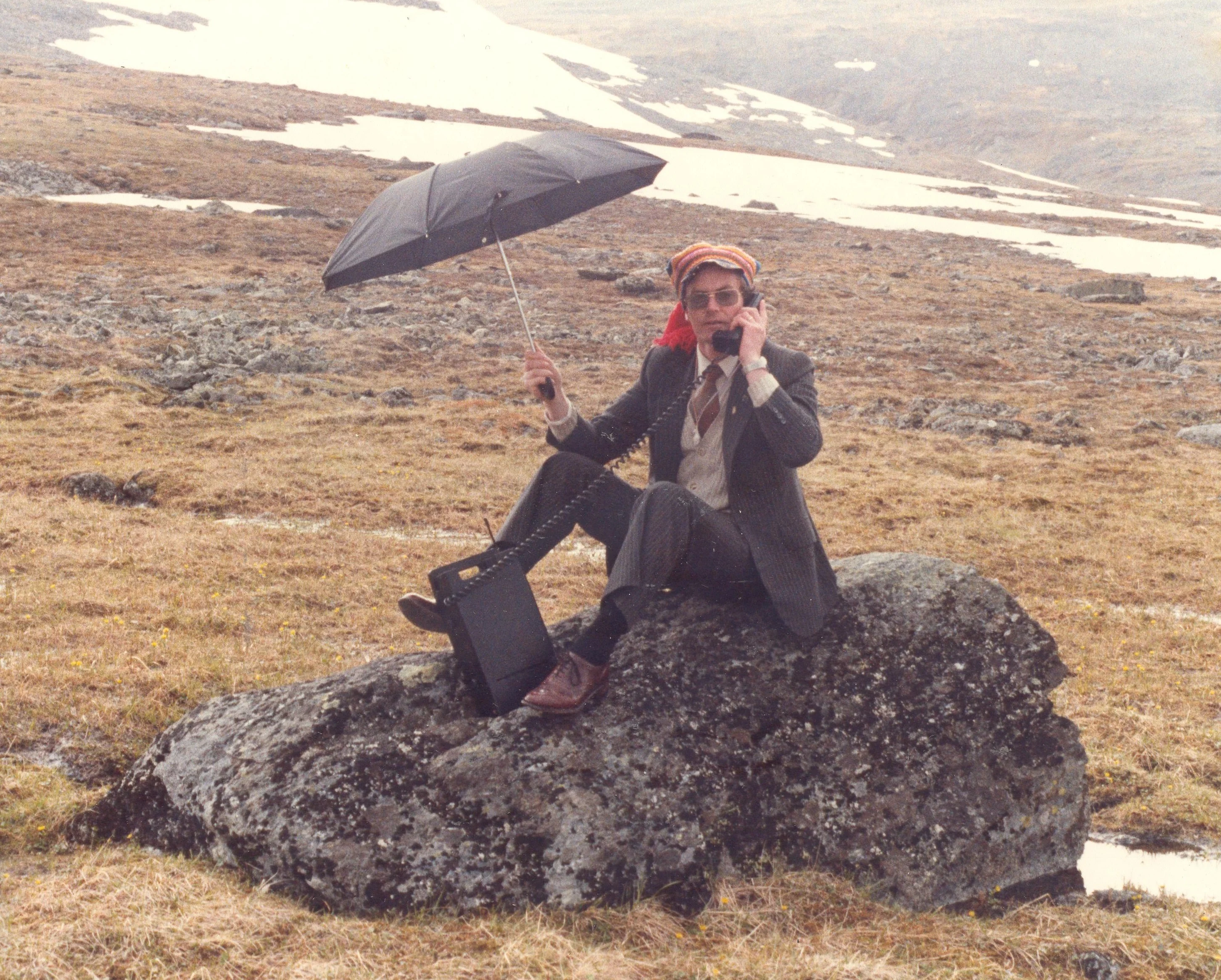 A person sitting on a rock holding an umbrella and talking on the phone