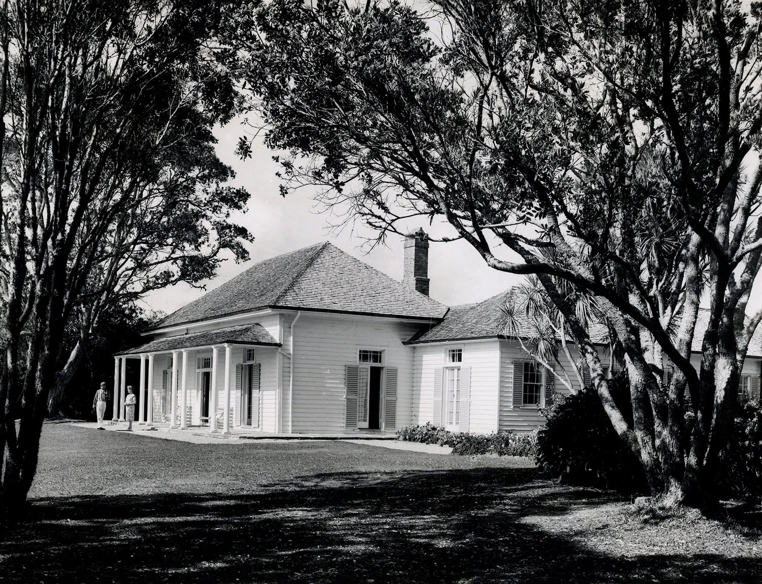 A white weatherboard house with a chimney and veranda, surrounded by grass and trees.