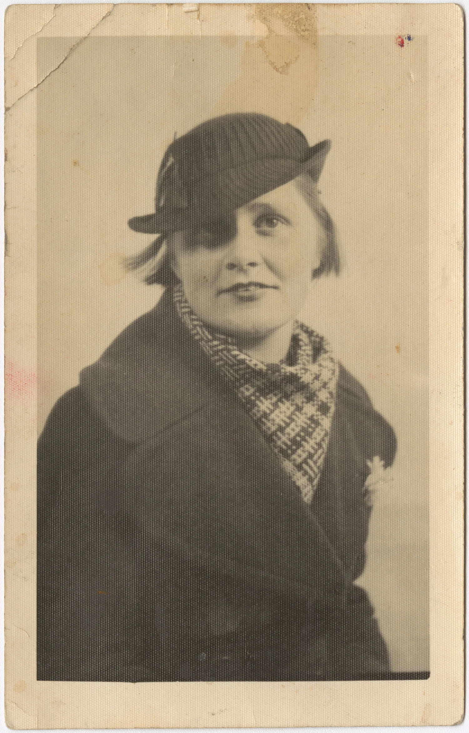 An old black and white photo of a woman wearing a hat, black jacket and a checkered scarf