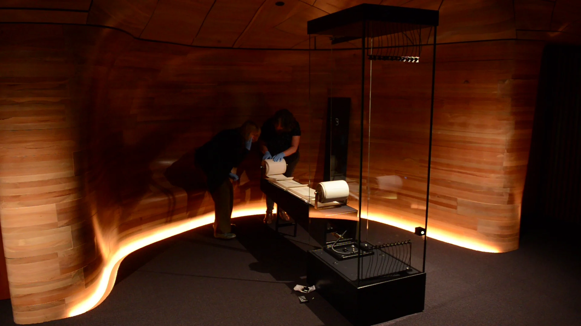 two women in a dark room, bending in front of a roll of paper with wooden circular wall behind