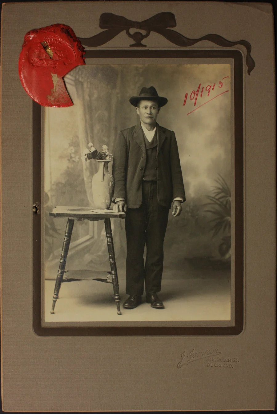Quan Hing stands posing for a portrait in a photography studio. He’s wearing a 3-piece suit and hat, and is next to a table with a vase of flowers on it.