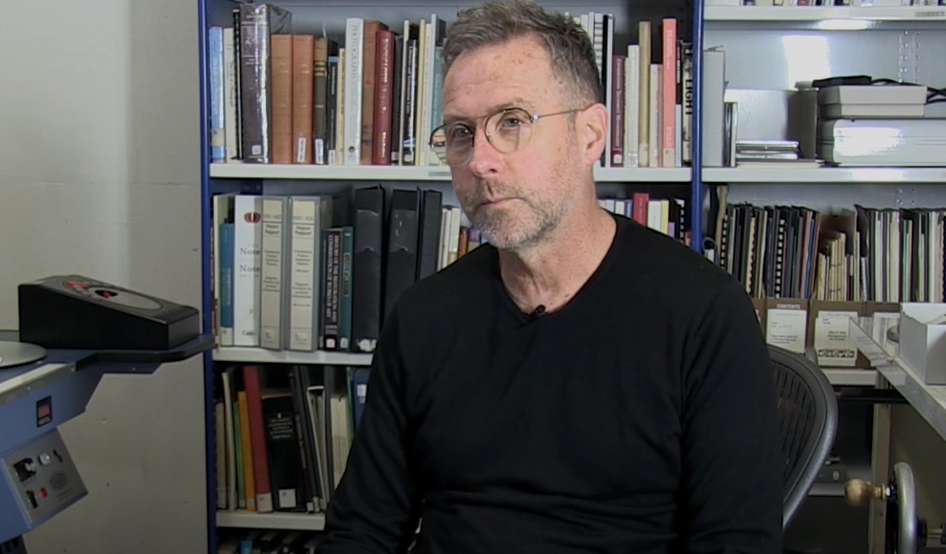 A man wearing black shirt and glasses, sitting in a chair, with a book shelf in the background 