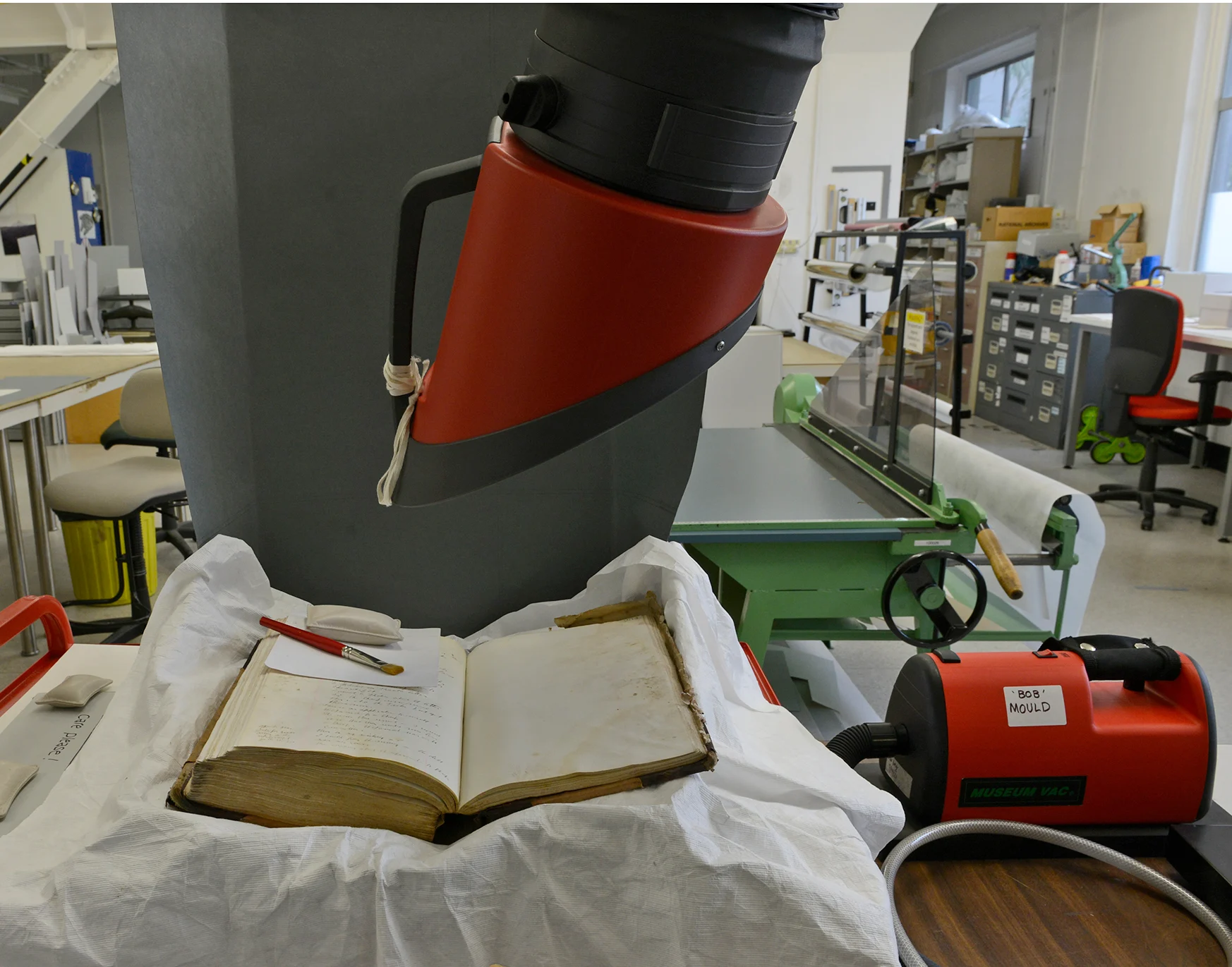An extraction hood and a museum vacuum cleaner over an old book. The book’s pages are yellowed with age and handwritten text. The background shows a typical lab setting with shelves stocked with various items and equipment.