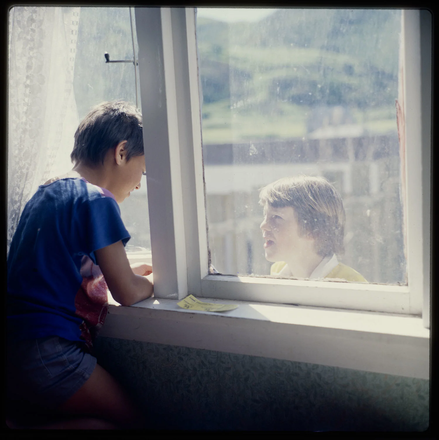 A young boy peeping out of a half open glass window and talking to another boy standing outside