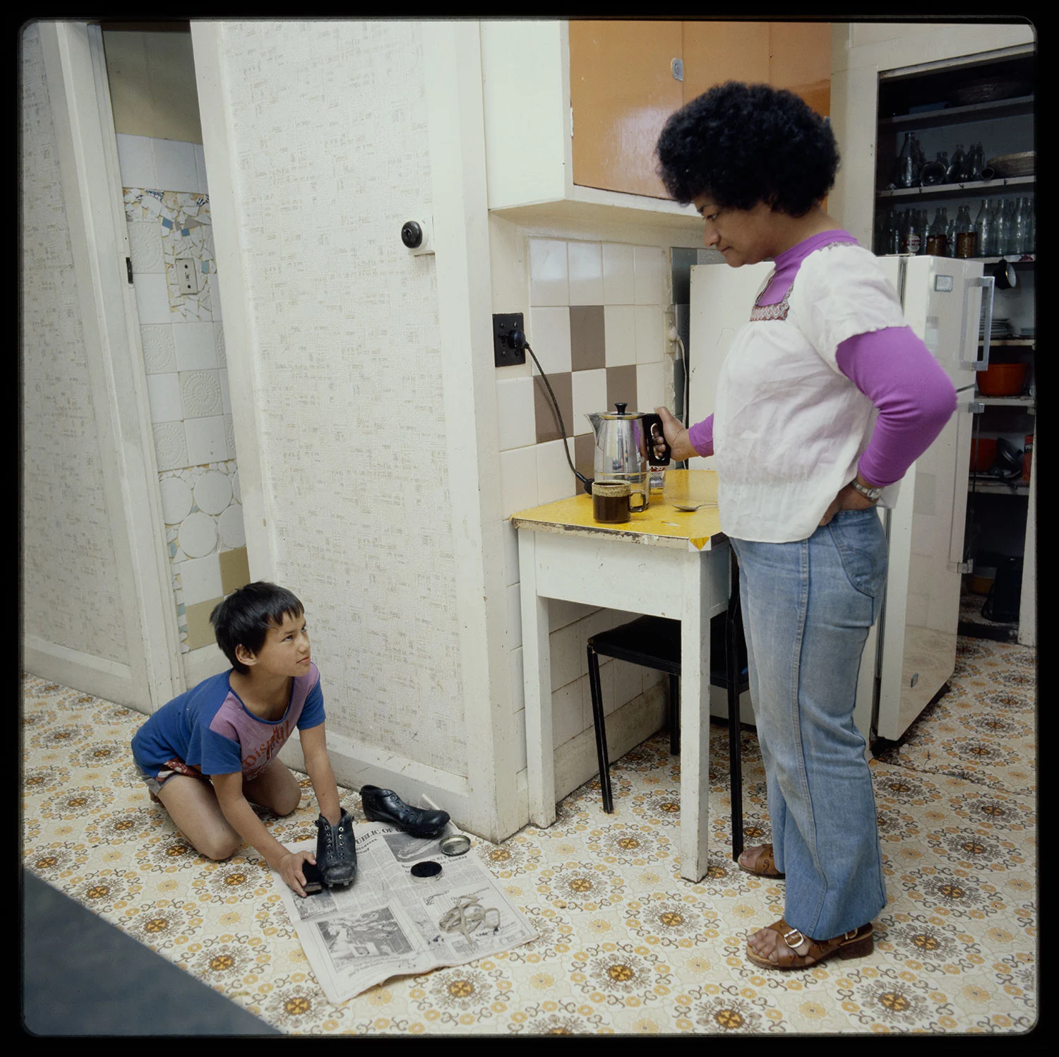 A boy wearing blue shirt is sitting on a kitchen floor polishing his black shoes and a woman with short hair and wearing shirt and pant standing in front of him