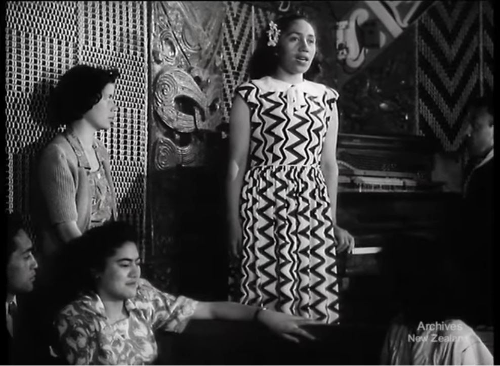 A still from the film 'Aroha'. A woman is standing and singing while others watch and listen.