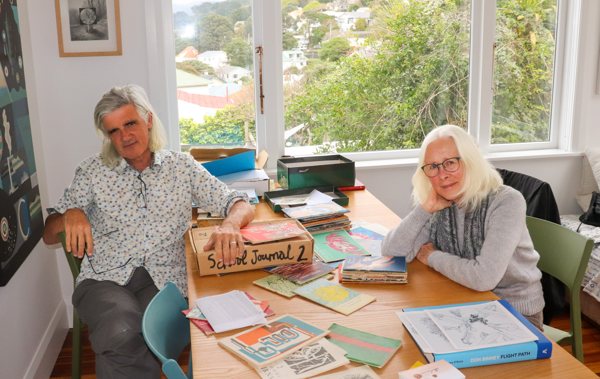 A seated grey haired man and seated grey haired female in a room at a table surrounded by books.