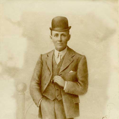 An old photo of a man wearing a suit and a hat standing in front of a pale yellow background, holding a pipe in his left hand and right hand in his pant's pocket
