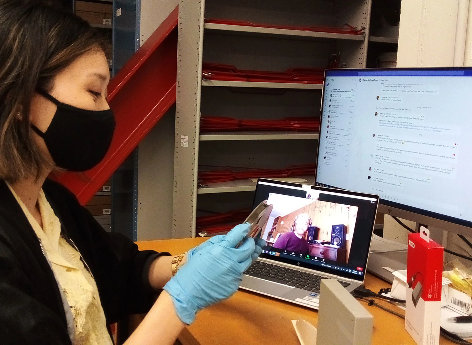 Side profile of a woman wearing a mask, sitting in front of the monitor and laptop holding a film reel