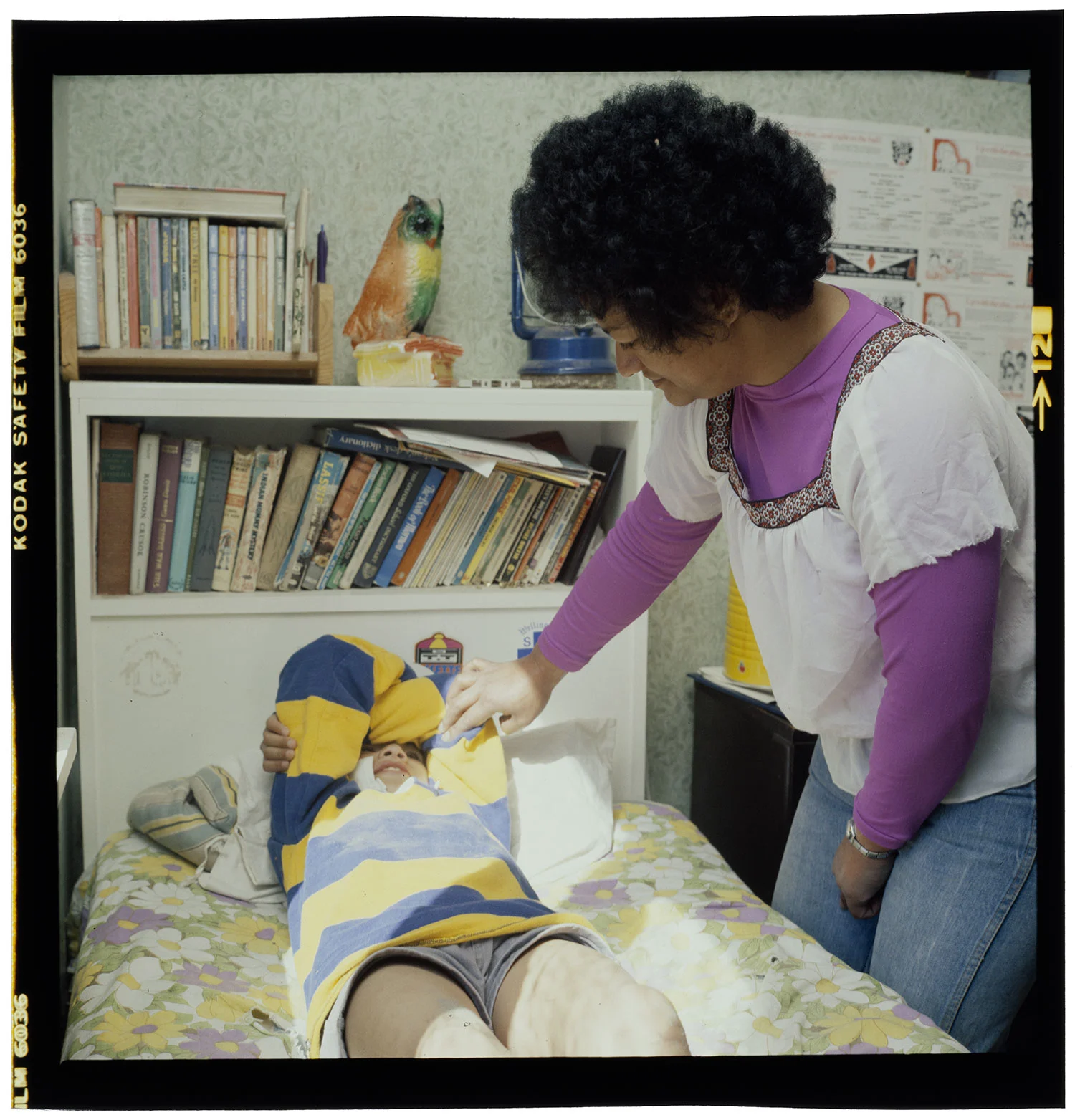 A woman with short hair wearing a purple full sleeves shirt and white top bending and touching a boy sleeping on a bed covering his face with his two hands
