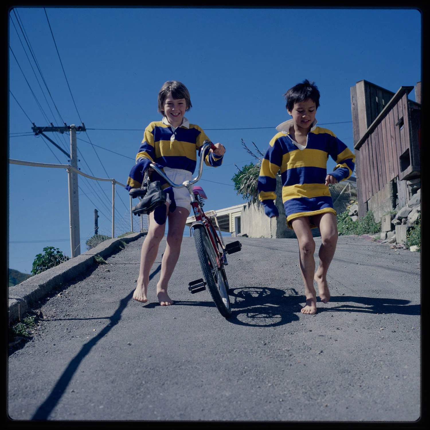 Two boys wearing blue and yellow striped shirts walking a road, one holding a bike in his hands 