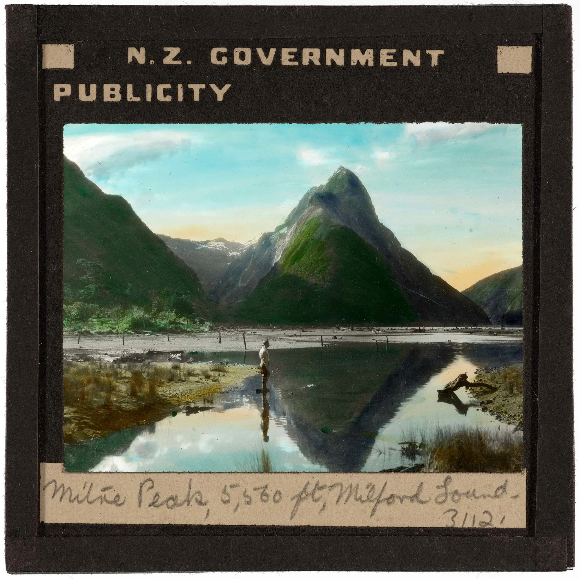 Craggy green mountains and a blue and orange sky dotted with clouds are reflected in water. A person is standing near the edge of the water.