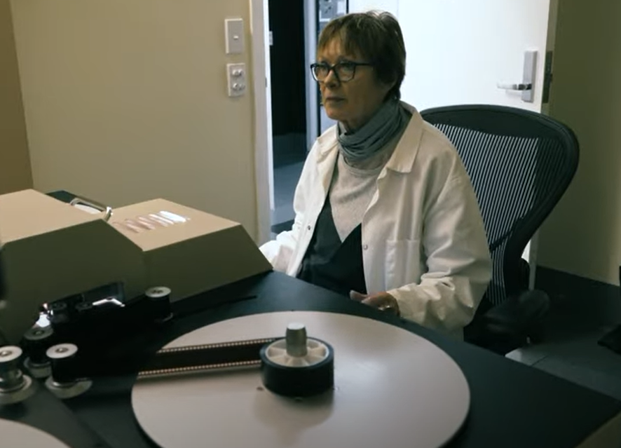 An old person with short hair wearing glasses and a white coat, sitting in a chair with a record disc in front