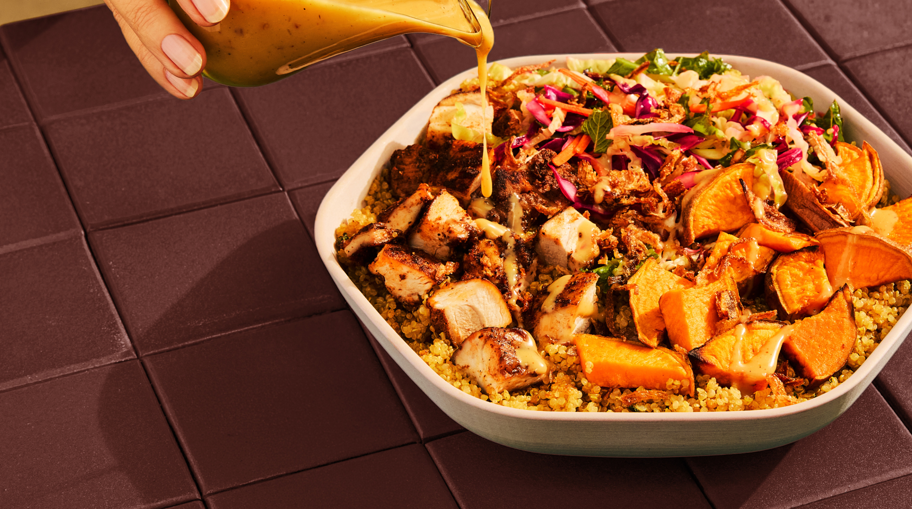 A hand pours golden dressing over a bowl of quinoa, grilled chicken, roasted sweet potatoes, and shredded cabbage slaw.
