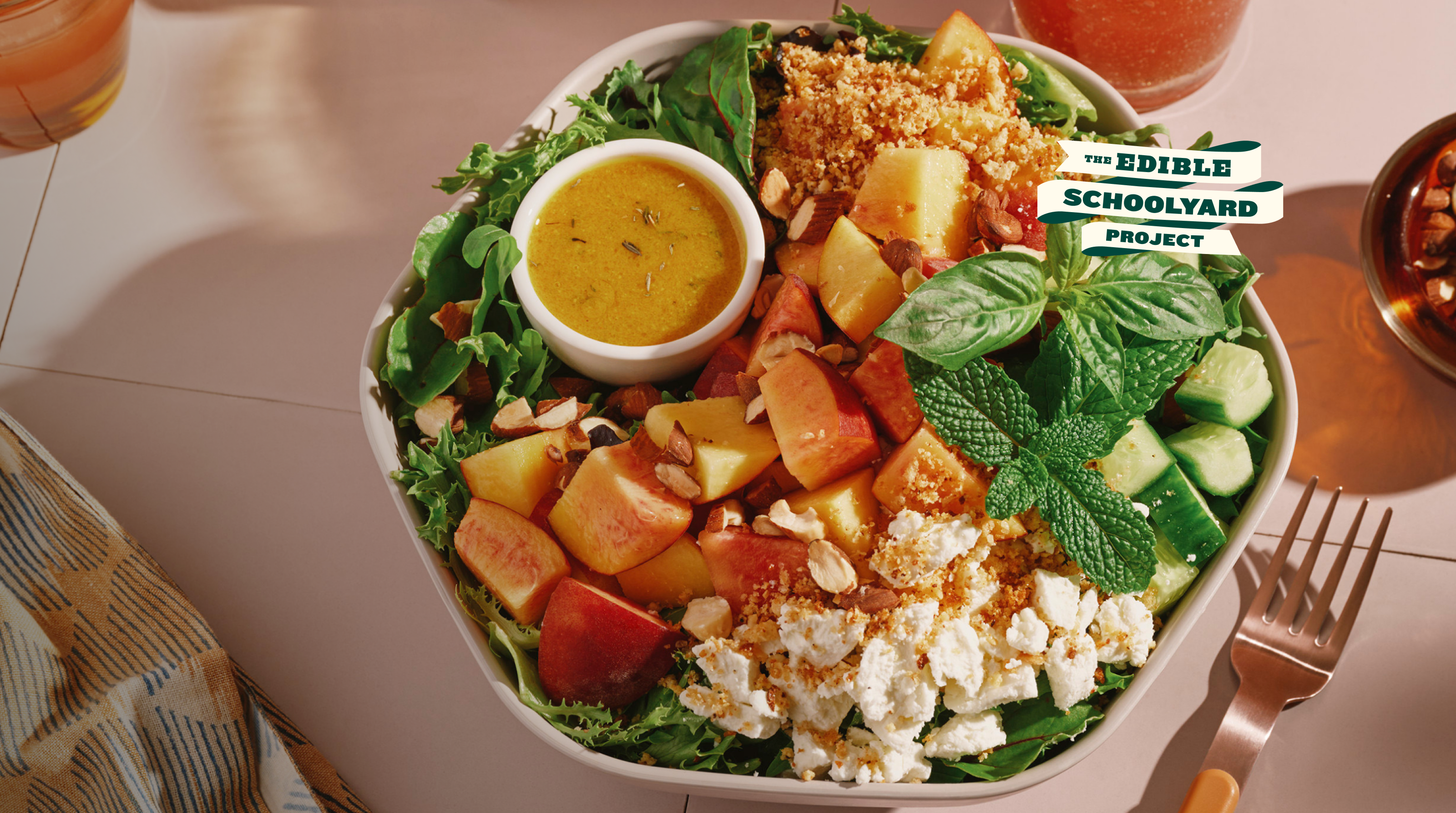  A white bowl holds a summer salad of arugula and spring mix topped with chunks of fresh peaches, crumbled goat cheese, cucumbers, sliced almonds, and garlic parm crunch, garnished with sprigs of fresh mint and basil, with a small dish of lemon tarragon vinaigrette nestled on the side. A copper fork rests on the light pink tiled surface beside the bowl, with a dish of chopped nuts and a beverage in the background. A logo reading "The Edible Schoolyard Project" appears in the upper left.