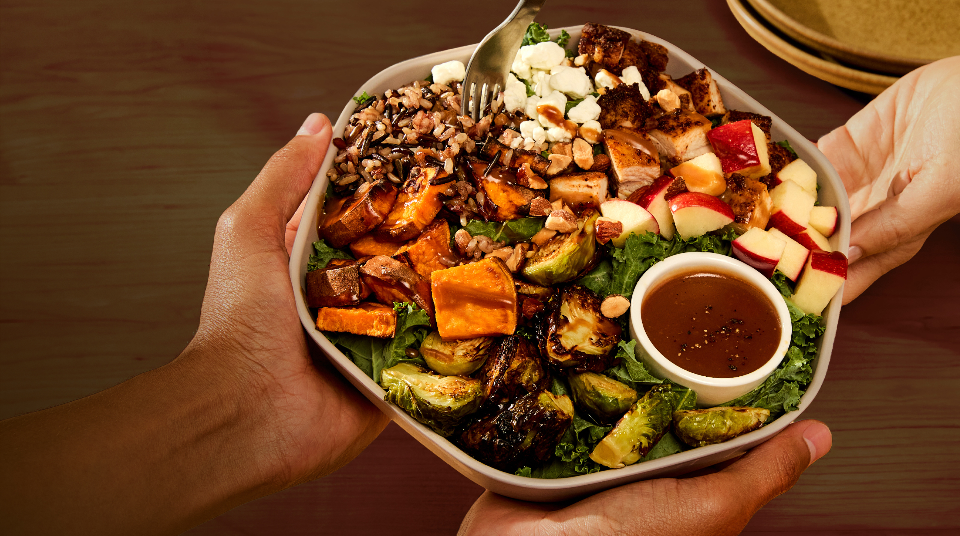 Overhead view of two hands presenting a square, light-colored bowl over a dark wooden table. The bowl holds a salad arranged in sections over a bed of kale: wild rice, caramelized roasted sweet potato wedges, charred Brussels sprouts, cubes of blackened chicken, crumbled goat cheese, diced red apples, and chopped almonds. A small ramekin of balsamic vinaigrette sits at the lower right, and a fork rests in the top of the bowl. Stacked plates are visible in the upper right corner.