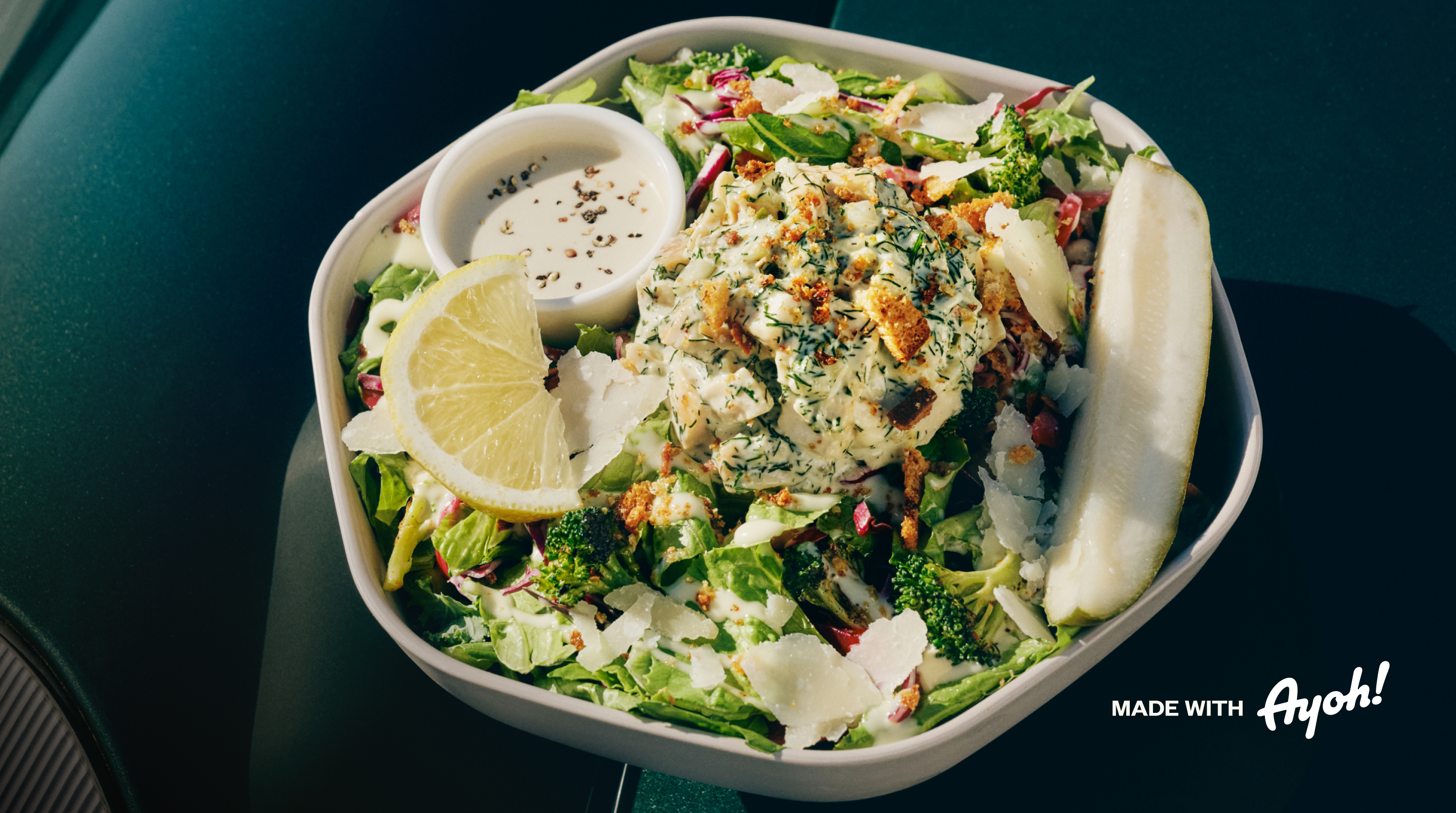 A hexagon white bowl of chopped salad on a dark green table: romaine, shredded red cabbage, roasted broccoli, and shaved parmesan, topped with a scoop of creamy chicken salad sprinkled with fresh dill and crispy breadcrumbs. A lemon wedge, a whole pickle spear, and a small cup of creamy dressing with cracked black pepper sit on the side.