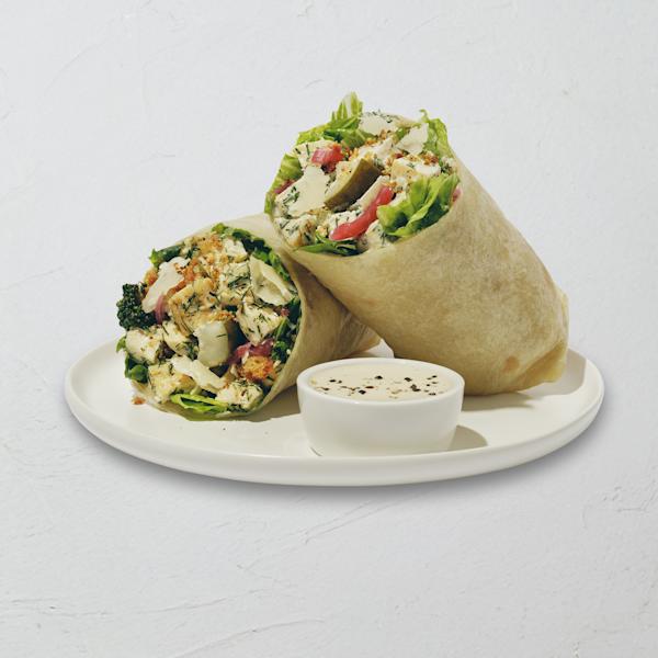 wrap cut in half on a white plate, showing a filling of chicken salad with fresh dill, romaine, pickles, pickled onions, shaved parmesan, roasted broccoli, and breadcrumbs, with a small cup of creamy dressing alongside.