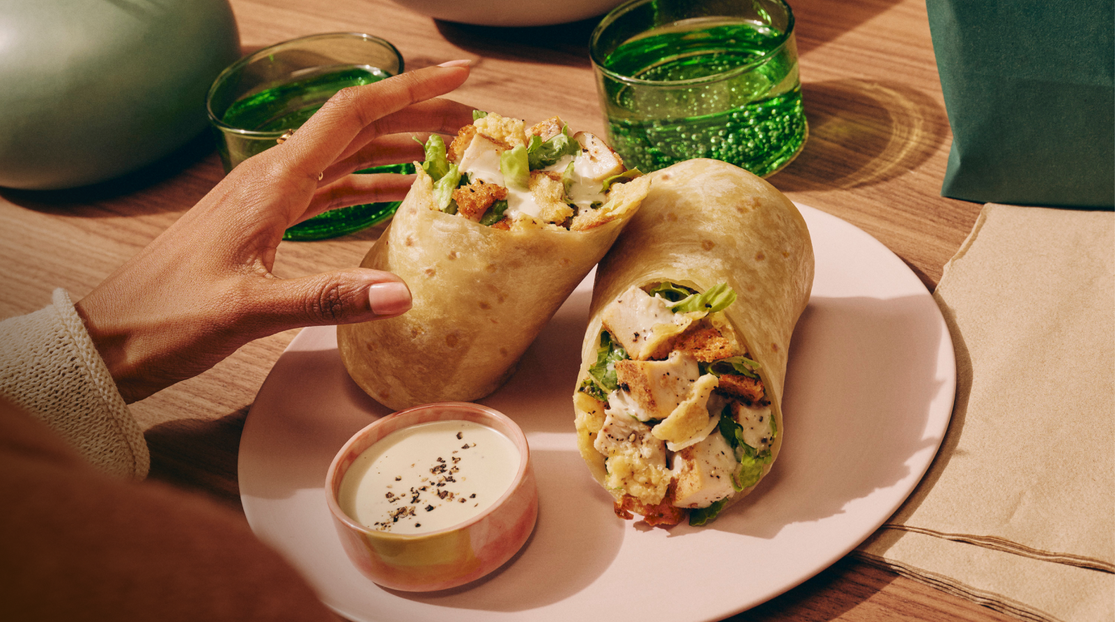 Hand picking up a Classic Chicken Caesar wrap on plate with caesar dressing on the side