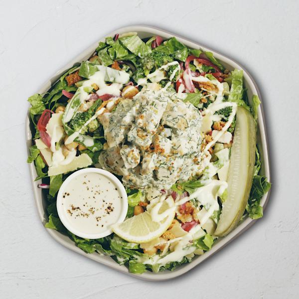 Overhead view of a salad bowl with a scoop of dill chicken salad in the center, surrounded by chopped romaine, roasted broccoli, chickpeas, pickled onions, shaved parmesan, and breadcrumbs, with a pickle spear, lemon wedge, and cup of creamy dressing on the side.