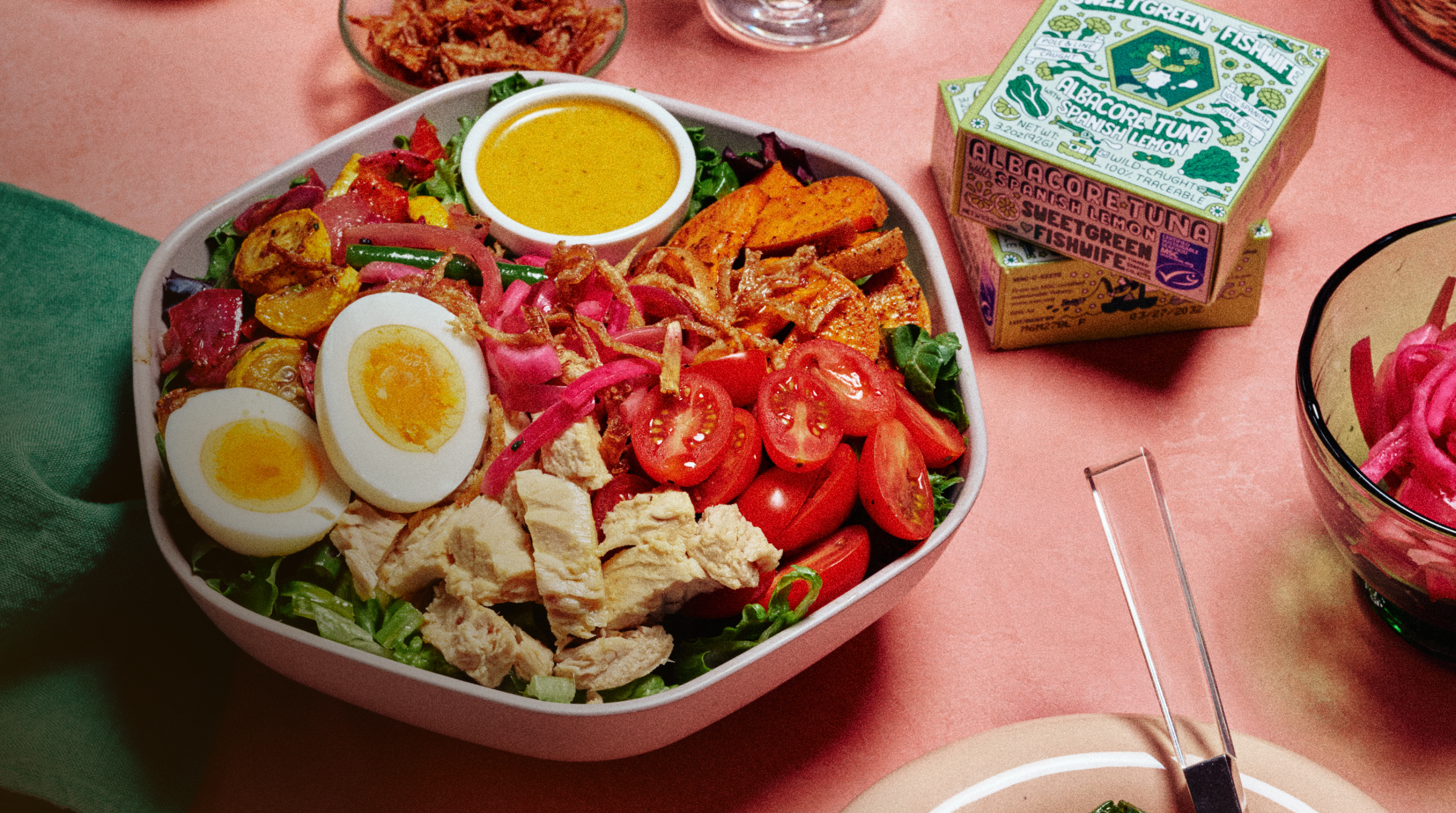 Overhead shot on a coral-pink surface: a light pink square bowl filled with mixed greens, chunks of albacore tuna, two halves of a soft-boiled egg with jammy yolks, halved cherry tomatoes, roasted sweet potato wedges, green beans, pickled red onion, and crispy fried shallots, with a small white ramekin of golden-yellow dressing nested in the middle. To the right, two boxes of sweetgreen × Fishwife "Albacore Tuna with Spanish Lemon" are stacked next to an open tin. Also in frame: whole lemons in a green mesh bag, a glass of water, a small bowl of extra crispy shallots, and a side plate of roasted vegetables.