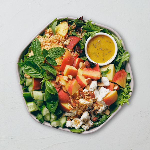 An overhead view of a summer salad in a white bowl on a light gray surface. Arugula and spring mix are topped with chunks of fresh peaches, crumbled goat cheese, chopped almonds, diced cucumbers, and golden garlic parm crunch, garnished with sprigs of fresh basil and mint. A small white dish of lemon tarragon vinaigrette flecked with herbs sits in the upper right.