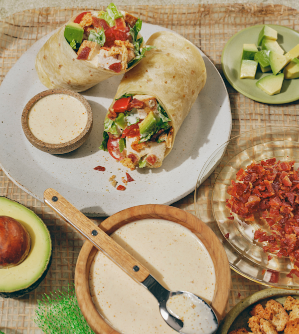 Cali chicken club wrap n a white plate, filled with lettuce, tomato, avocado, and bacon bits, served with a small bowl of creamy dressing. Surrounding it are ingredient dishes — diced avocado, crumbled bacon, a halved avocado, and a wooden bowl of dressing with a spoon.