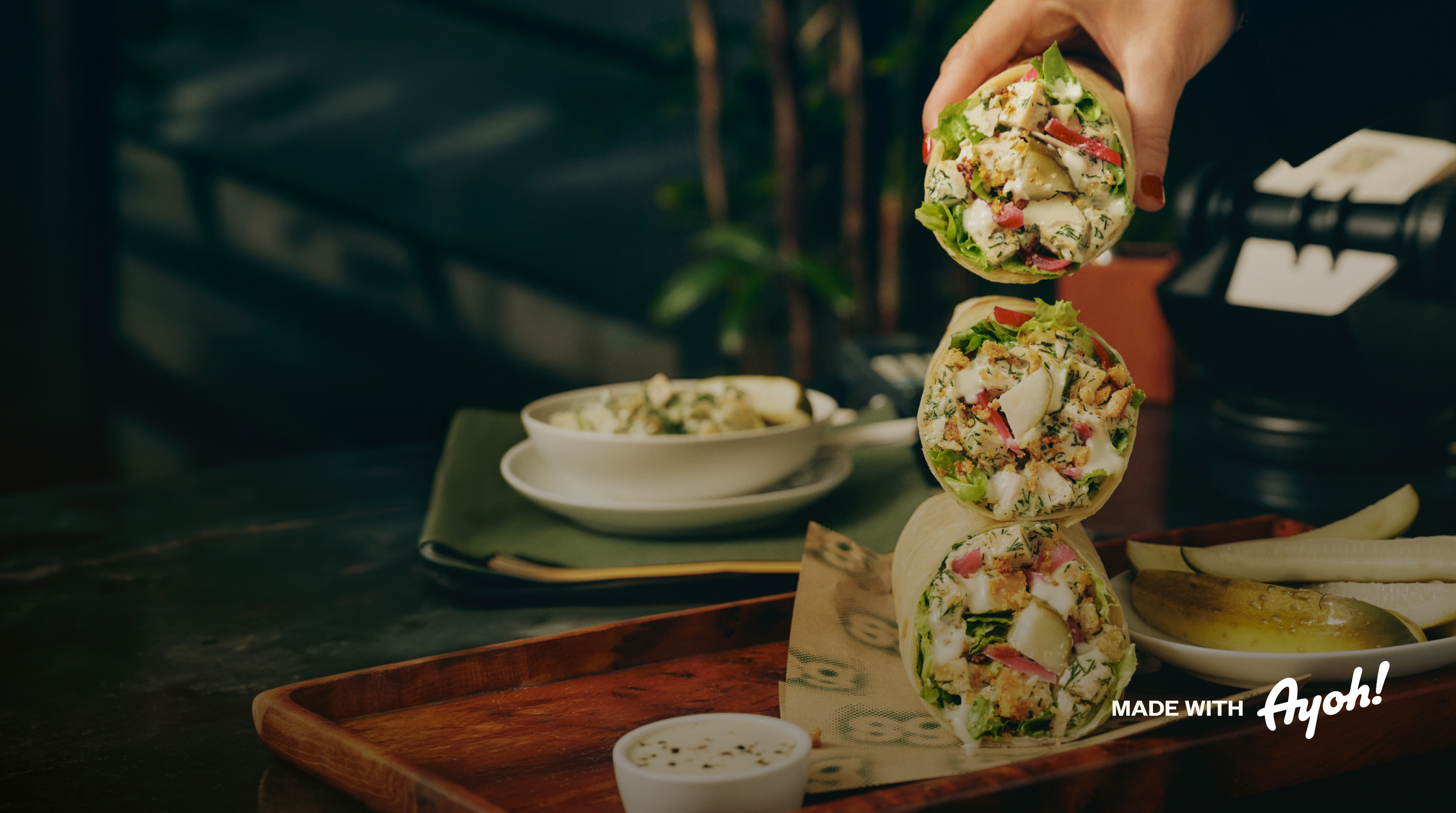 Three halves of a chicken Caesar-style wrap stacked vertically, held at the top by a hand with red nail polish, showing a filling of chopped chicken, romaine, dill, pickles, pickled red onion, and crunchy breadcrumbs in a creamy dressing. 