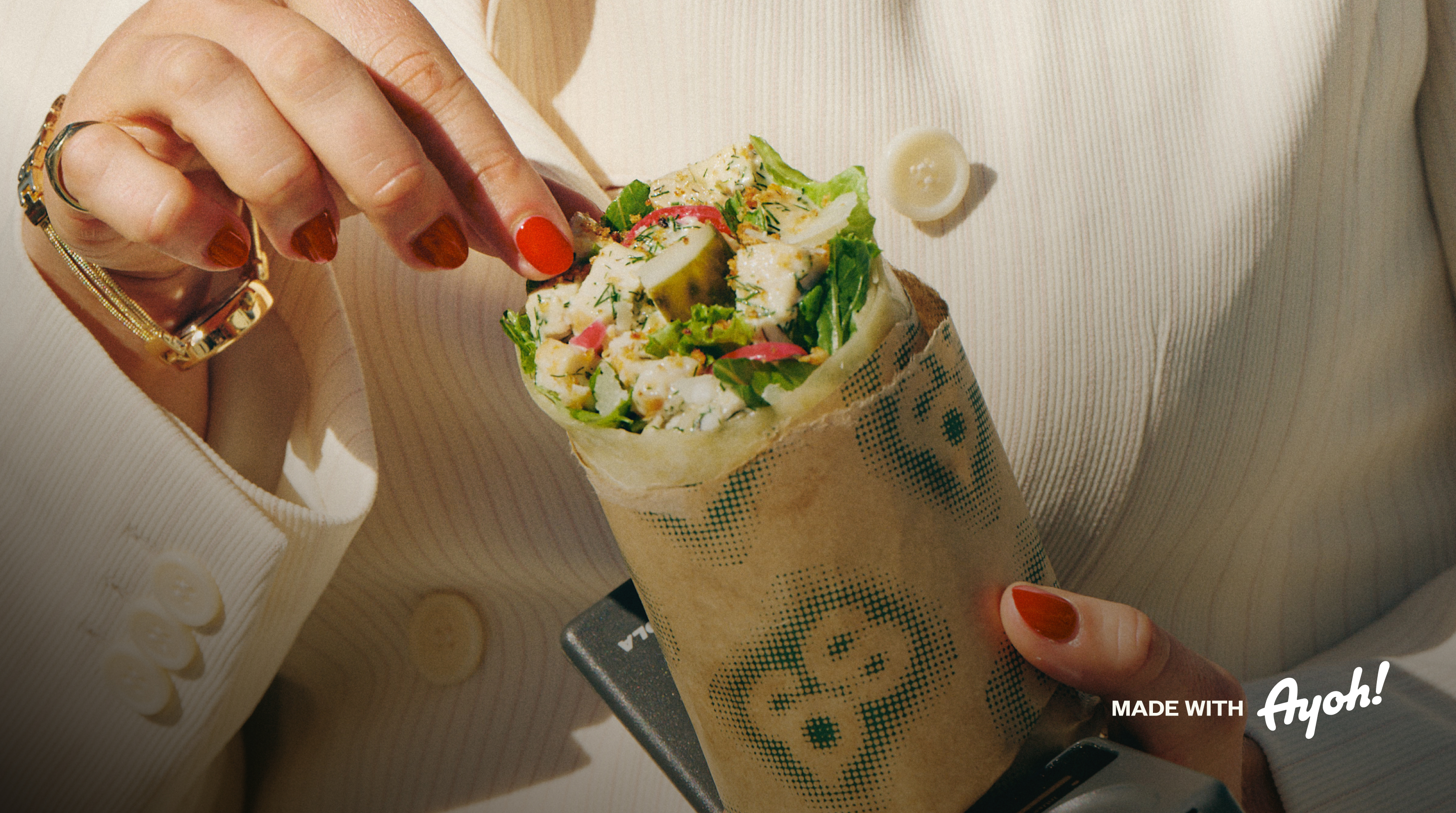 A person in a cream ribbed knit cardigan holds a wrap in a kraft paper sleeve printed with a green Sweetgreen pattern. One hand with red-painted nails reaches toward the open top, which is filled with chopped chicken, romaine, pickle slices, pickled red onion and fresh dill. Text on the right reads “Made with Ayoh!”