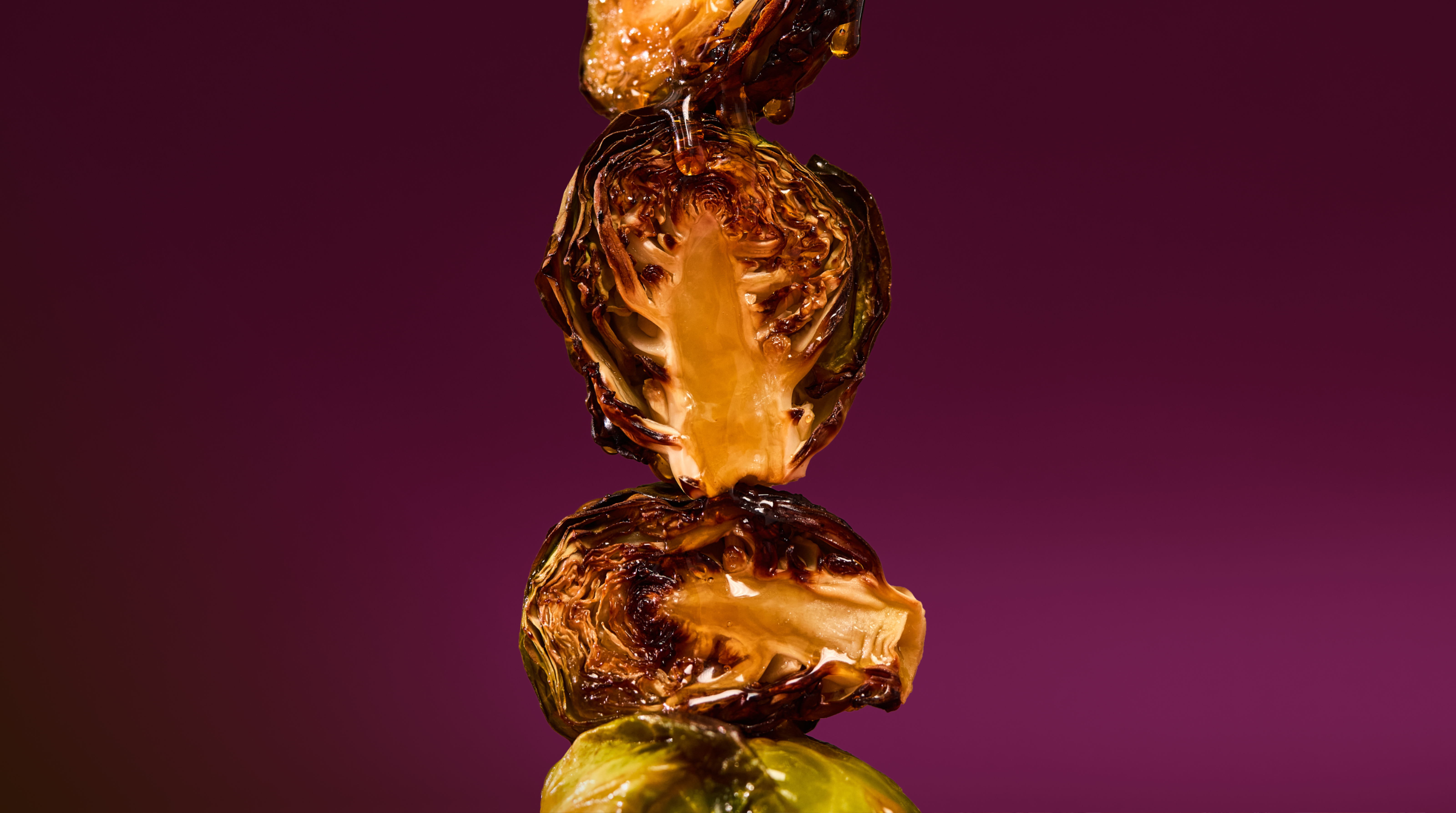 Vertical stack of caramelized, maple-glazed Sweetgreen Brussels sprout halves dripping with glaze against a deep magenta background.