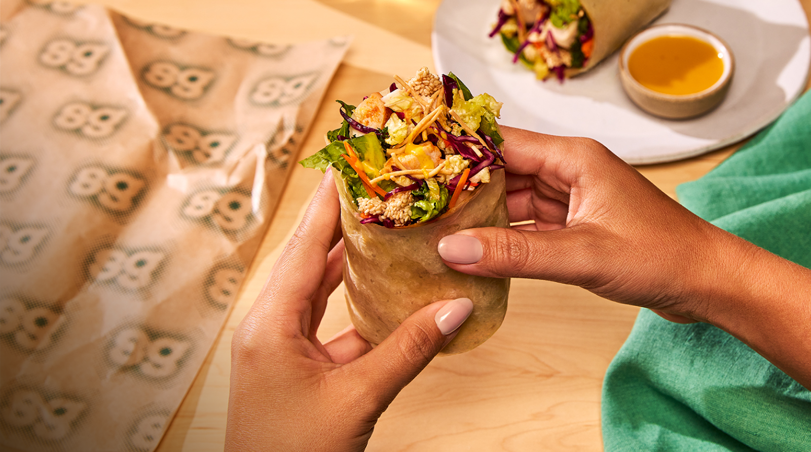 Wide crop on two hands holding a Chicken Sesame Crunch Wrap upright over a light wood table. The open top reveals shredded purple cabbage, romaine, carrots, grilled chicken, sesame seeds, crispy wontons, and yellow-orange dressing. A folded sweetgreen-branded paper bag fills the left third of the frame. In the upper right, partially cropped, a white plate holds a second wrap beside a small stoneware cup of the same dressing. A green cloth napkin sits at the right edge.