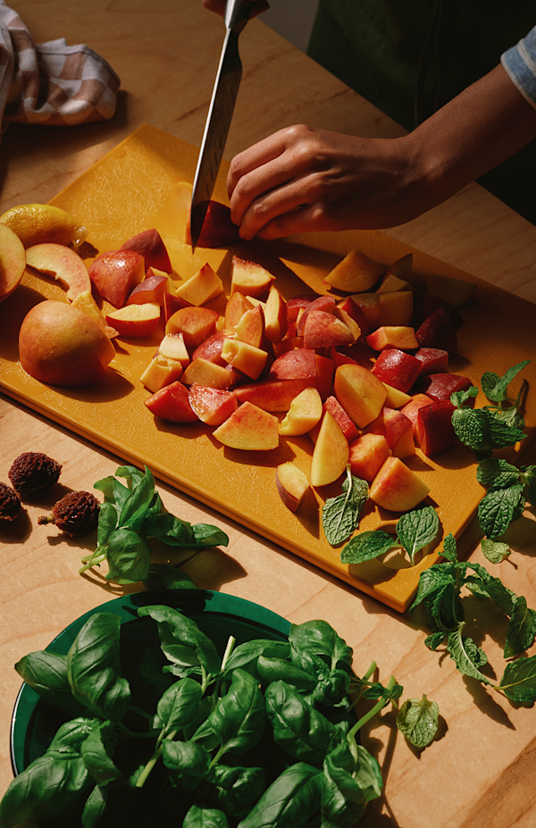 A chef uses a knife to cut peaches on a cutting board. Spring of mint and basil surround the cut up peaches