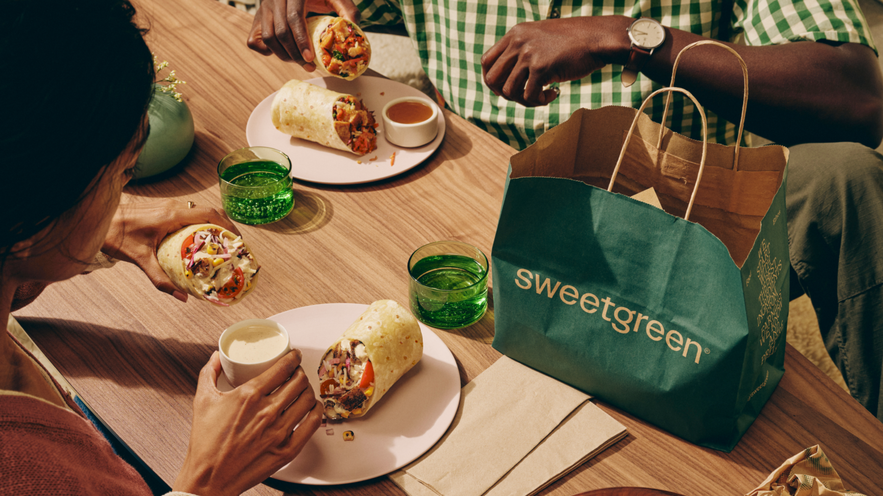 Two people at a wood table sharing a Sweetgreen takeout meal — wraps on plates with a side of sauce, glasses of water, and a dark green Sweetgreen bag with tan handles sitting on the table.