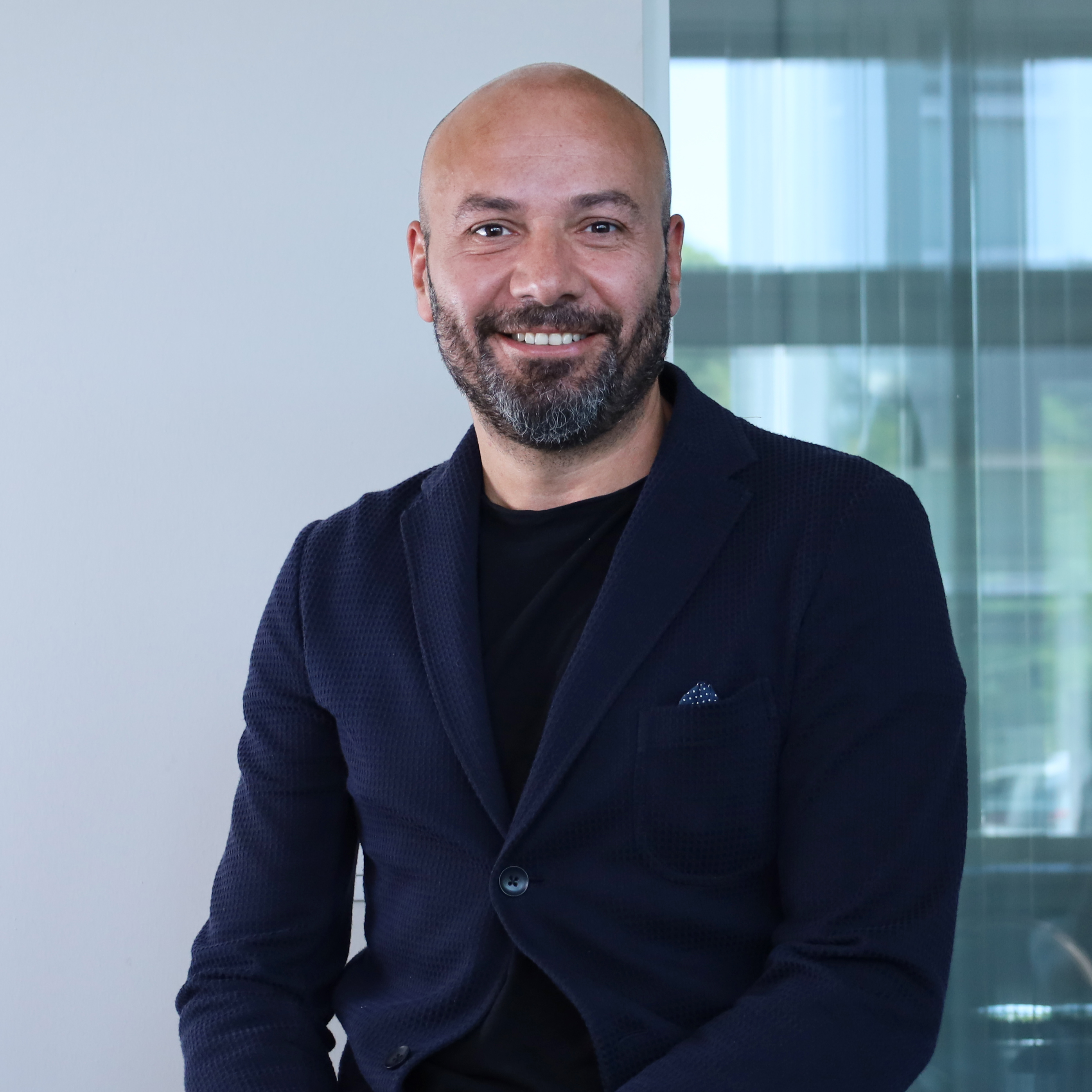 Ritratto a mezzo busto di un uomo sorridente e con la barba ed il Chief Operating Officer di Redify. Indossa una giacca blu scuro sopra una maglia nera. Lo sfondo è luminoso e sfocato, con una vetrata che suggerisce un ambiente d'ufficio