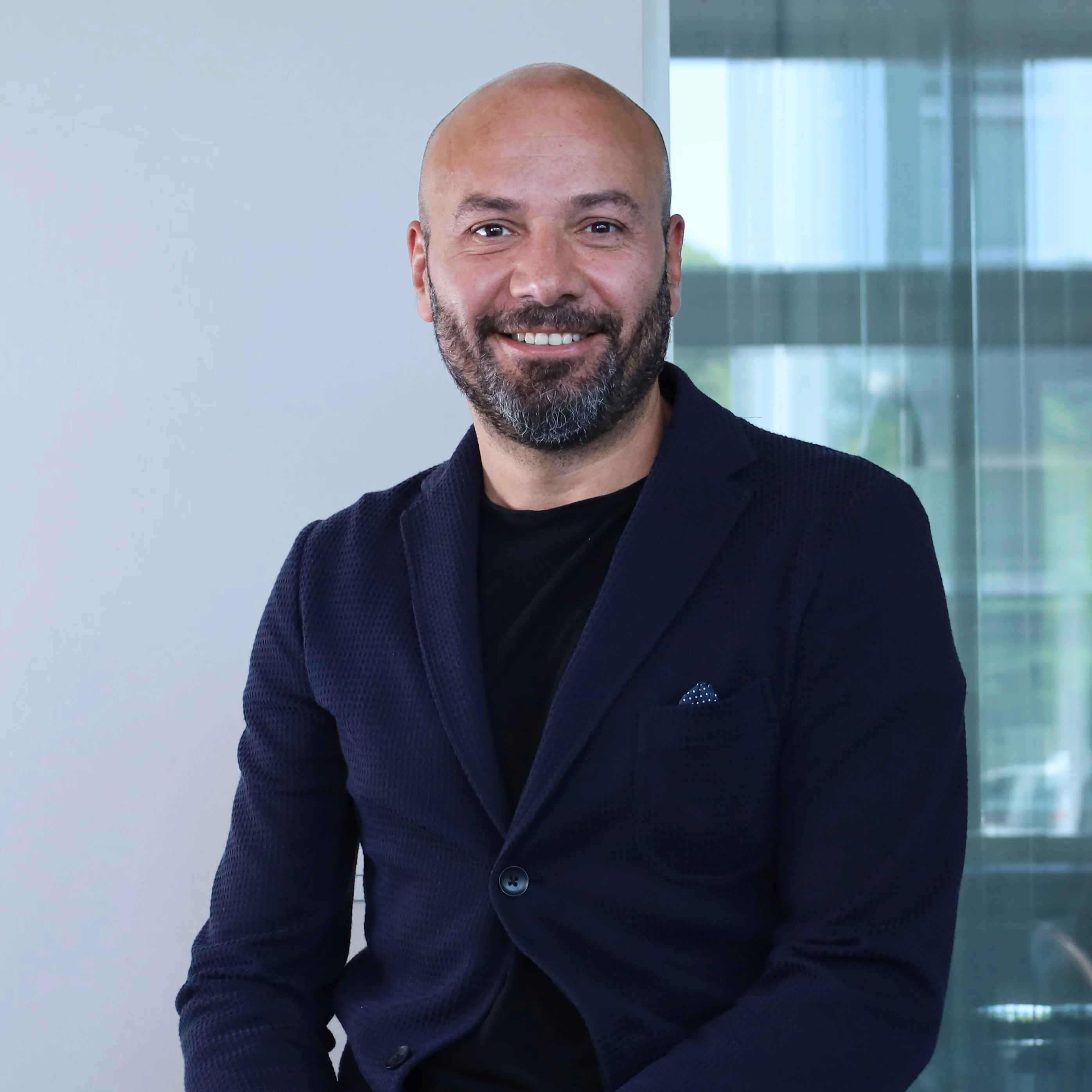 Ritratto a mezzo busto di un uomo sorridente e con la barba ed il Chief Operating Officer di Redify. Indossa una giacca blu scuro sopra una maglia nera. Lo sfondo è luminoso e sfocato, con una vetrata che suggerisce un ambiente d'ufficio